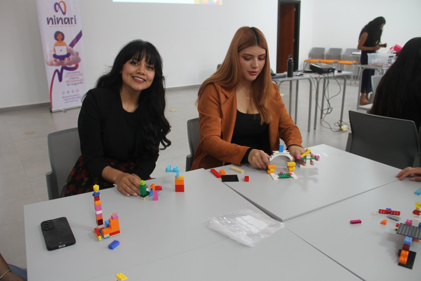Dos participantes construyen modelos con bloques en el taller de marca personal de Ninari, junto al banner de la organización